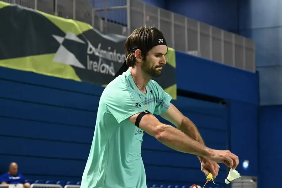 Lucas Mazur au service, volant et raquette en main, devant une banderole Badminton Ireland